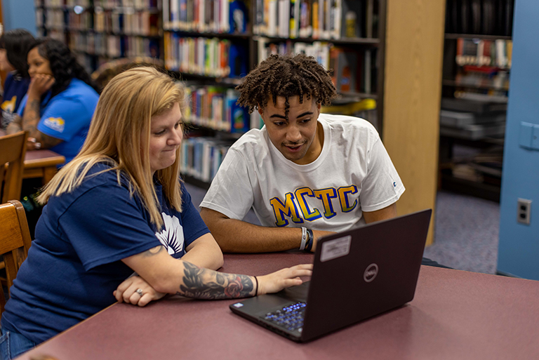 student and staff member in a library and working on a laptop