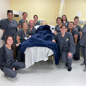 Nursing students in gray uniforms around a hospital bed with medical mannequin.