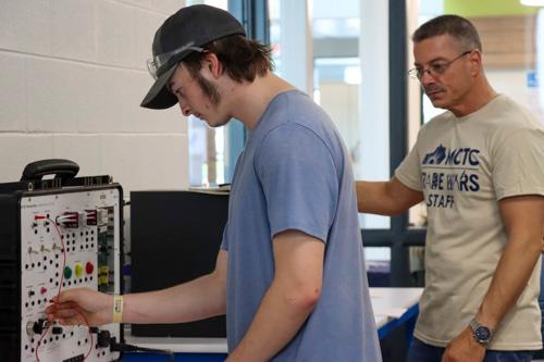 student working on a circuit board with a Trade Wars staff member watching