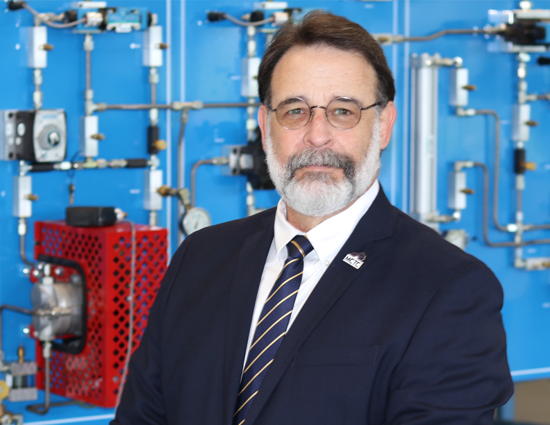Interim President Russ Ward standing in front of a blue mechanical background in a technical lab.