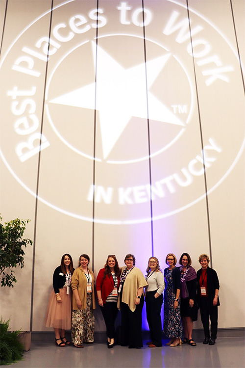 Group of eight employees standing under the Best Places to Work in KY logo projected onto the wall.