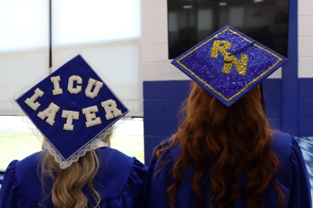 two grads with decorated caps; one that says "ICU Later" and one that says "RN"