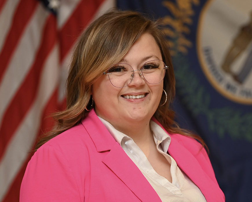 Kaitlin Stewart wearing a pink blazer, standing in front of a United States flag and a Kentucky state flag.