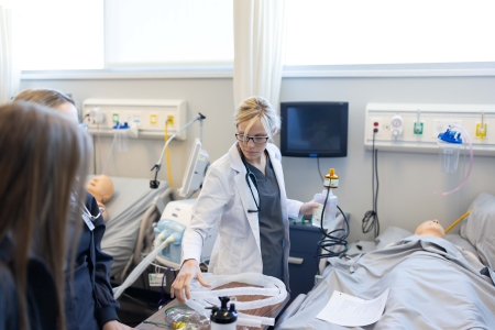 instructor and students by a bed with a mannequin in the respiratory lab